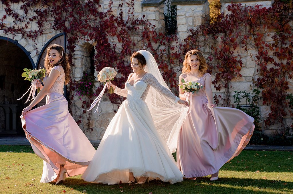 atrakcyjna-usmiechnieta-panna-mloda-i-druhny-tancza-i-bawia-sie-przed-kamiennym-budynkiem-pokrytym-czerwonym-bluszczem